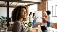 Professional African American businesswoman with curly hair smiling in modern office with diverse team collaborating in background during meeting.