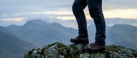 Hiker in dark pants and boots standing on rocky mountain peak with vast alpine landscape and distant ranges in background, representing outdoor adventure and achievement.