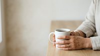 Mature woman with medium skin tone holding warm beverage in ceramic mug at wooden table, wearing casual cream sweater, relaxed lifestyle moment for wellness and comfort themes.