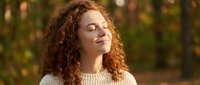 Young mixed-race woman with curly hair enjoying peaceful moment outdoors in warm sunlight, eyes closed in serene expression wearing cream sweater.