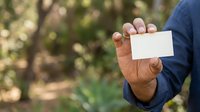 Male professional with dark skin tone holding blank white business card outdoors in natural garden setting, suitable for corporate branding, networking, and business identity projects.