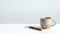 Minimalist ceramic coffee cup with copper accent and wooden pen on clean white desk against neutral background for modern workspace design.