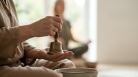 Hands holding traditional brass bell during meditation practice in peaceful home setting with natural light creating calm spiritual atmosphere for mindfulness.