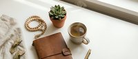 Cozy workspace flat lay with leather notebook, coffee cup, succulent plant and wooden beads on white desk surface for productivity and lifestyle content.