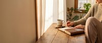 Woman in beige sweater writing in notebook at wooden table by window with warm sunlight and coffee cup creating cozy morning atmosphere for journaling.