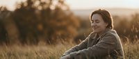 Peaceful middle-aged Caucasian woman sitting in golden autumn field during sunset, enjoying quiet moment in nature with warm light and serene expression.