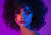Young African American woman with natural curly afro hair in vibrant neon purple and blue lighting, dramatic beauty portrait showing glowing skin and expressive eyes.
