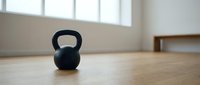 Black kettlebell on wooden floor in bright gym studio with windows, ready for strength training workout and fitness exercise routines.