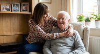 Middle-aged Caucasian woman embracing elderly Caucasian man on sofa in cozy living room with family photos, showing intergenerational love and care at home.