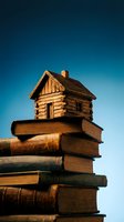 Miniature wooden log cabin sitting on stack of old vintage books against blue sky background symbolizing knowledge, education and learning foundation.
