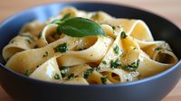 Fresh pappardelle pasta with herbs and olive oil in dark ceramic bowl, garnished with basil leaves, close up view of traditional Italian cuisine.