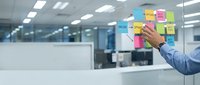 Businessman in blue shirt placing colorful sticky notes on glass wall in modern office space for project planning and brainstorming sessions.