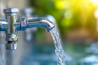 Chrome water tap with flowing crystal clear water stream against blurred green natural background, close-up shot with soft bokeh effect and sunlight.