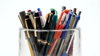 Assorted pens and markers in clear glass container, office supplies for writing, drawing, and design work on white background.