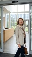 Professional middle-aged Caucasian businesswoman in beige blazer standing at modern office entrance with glass doors and contemporary interior design.