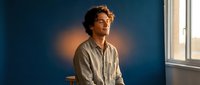 Young handsome man with curly hair sitting by window in warm sunlight, contemplative mood, casual shirt, peaceful morning atmosphere for lifestyle content.