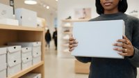 Young African American woman holding blank white tablet in modern retail store environment with shelving and customers in background.