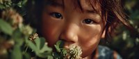 Young Asian girl with messy hair hiding behind green clover flowers in natural outdoor setting, childhood innocence and nature connection concept.
