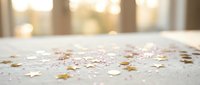 Golden confetti stars scattered on white wooden surface with warm bokeh lights creating festive celebration atmosphere for party decorations.