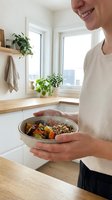 Young Caucasian man holding bowl with roasted vegetables and quinoa in modern kitchen with potted plants and bright windows.