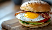 Breakfast sandwich with sunny side up egg, crispy bacon strips, fresh lettuce and avocado slices on toasted brioche bun served on wooden cutting board.