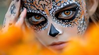 Creative sugar skull makeup with orange and silver glitter details on pale skin, dramatic eye design and black nose accent against blurred orange background.