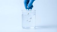 Gloved hand dropping white tablets into a glass beaker with water, showing dissolution process in a laboratory setting.