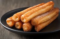 Fresh churros dusted with sugar served on black plate against wooden background, close up view of traditional Spanish pastry dessert.