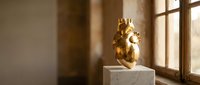 Golden anatomical heart sculpture displayed on white marble pedestal in sunlit room with warm natural lighting and wooden window frame.
