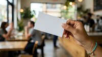 Hand holding blank white card in modern office environment with blurred colleagues and plants in background, suitable for business branding and corporate presentations.
