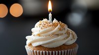 Single vanilla cupcake with white frosting swirl topped with lit birthday candle and gold sprinkles on dark background with soft bokeh lights.