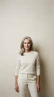 Confident mature Caucasian woman with gray hair wearing cream sweater and pants standing against neutral beige wall background for lifestyle content.