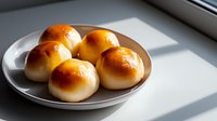 Fresh homemade baked buns with golden brown crust served on ceramic plate in natural window light creating dramatic shadows on white surface.