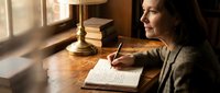 Mature Caucasian woman writing in journal at wooden desk by window, warm lamp light, thoughtful mood, cozy home office setting.
