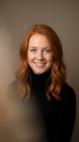 Professional portrait of smiling young Caucasian woman with wavy red hair wearing black turtleneck sweater against neutral beige background.