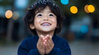 Young Asian boy with curly black hair wearing navy sweater smiles joyfully with hands clasped, against blurred bokeh lights background creating magical holiday atmosphere.