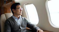 Middle-aged Asian male businessman wearing glasses and casual cardigan looking out airplane window during flight, travel and business concept.