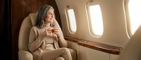 Elegant mature businesswoman enjoying coffee in luxury private jet cabin during flight with warm sunlight streaming through windows.