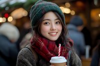 Young Asian woman wearing winter knit hat and scarf holding coffee cup outdoors with festive bokeh lights in background, expressing genuine joy and warmth.