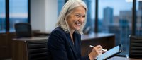 Senior Caucasian businesswoman with gray hair smiling while writing notes in modern office with city skyline view. Professional executive planning strategy.