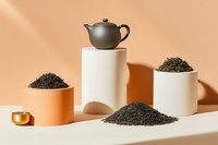 Minimalist tea composition with clay teapot on geometric podiums, loose black tea leaves scattered and in ceramic bowls against peachy background with dramatic shadows.