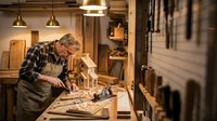 Senior craftsman woodworker in plaid shirt using hand plane on workbench in traditional workshop with hanging tools and warm lighting atmosphere.