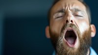 Bearded man yawning with eyes closed against dark background, showing natural tiredness and fatigue in dramatic lighting, expressing exhaustion and need for sleep.