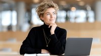 Professional blonde businesswoman in black sweater smiling thoughtfully at modern office workspace with laptop for corporate presentations and marketing.
