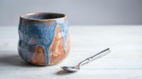 Handmade ceramic mug with blue and orange glaze drips on wooden surface with vintage spoon for coffee tea drinking and kitchen decor.