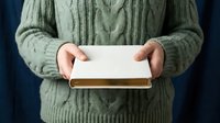 Person in green cable knit sweater holding white gift box with gold ribbon against dark blue background for holiday presents and special occasions.