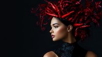 Young mixed race woman wearing dramatic red feather headdress and black high neck dress against dark background, profile view showcasing artistic fashion styling.