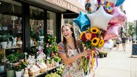 Young woman with long brown hair and fair complexion wearing a colorful floral dress, holding a vibrant bouquet of sunflowers and mixed flowers outside a flower shop storefront with balloons.