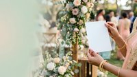 Woman with medium skin tone holding blank white card at floral wedding ceremony backdrop with pink roses and greenery, suitable for invitations, greetings, announcements.
