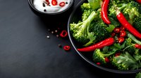 Fresh broccoli and red chili peppers in black pan with yogurt sauce on dark background for healthy cooking and spicy vegetarian meal preparation.